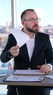 İş yerindeki sinirli iş adamı. Takım elbiseli gergin bir adam çalışma yerinde oturup kağıtları fırlatıyor. İşadamı ofisten biriyle öfkeyle konuşuyor. Dikey video
