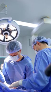 Üç erkek cerrah birlikte iş birliği yaparak ameliyat yapıyor. Doktorlar ameliyathanede yuvarlak lambanın altında çalışır. Dikey video.