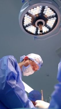 Erkek cerrahi ekibi bir hastayı ameliyat ediyor. Doktorlar modern ameliyathanede yuvarlak lamba altında çalışırlar. Düşük açı. Dikey video.