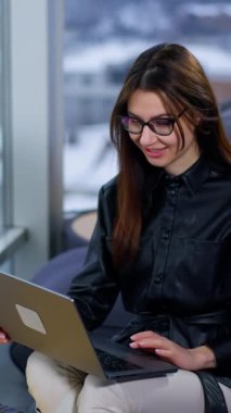 Ofiste dizüstü bilgisayarla çalışan kadın girişimci. Gözlüklü kadın ekrana bakıp gülümsüyor ve uzun siyah saçlarına dokunuyor. Dikey video