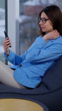 Gevşemiş esmer koltukta oturmuş telefona bakıyor. Büyük pencerenin yanındaki rahat koltukta aletini kullanan bir kadın. Dikey video