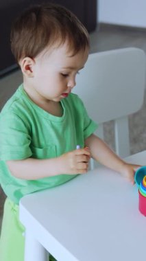 Masanın yanında yeşil tişörtlü güzel bebek oyuncağı duruyor. Küçük çocuk oyuncaklara odaklanmış. Üst Manzara. Dikey video