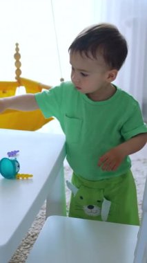 Beyaz bir çocuk masanın yanındaki odada dikiliyordu. Güzel çocuk parlak oyuncaklarla oynuyor. Dikey video