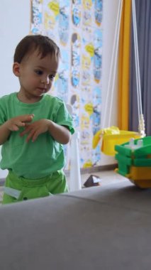 Yeşil elbiseli küçük güzel çocuk odada oynuyor. Küçük çocuk kanepenin yanında giden trene bakıyor. Evet. Dikey video
