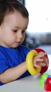 Çekici, beyaz bir erkek bebek masada piramitlerle oynuyor. Yeni yürümeye başlayan çocuk renkli yüzükleri eline takar ve neşeli bir şekilde alkışlar. Dikey video