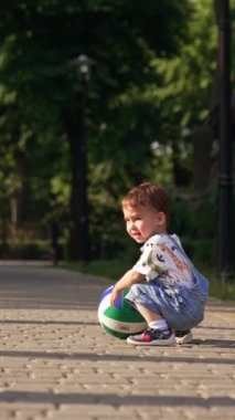 Küçük çocuk topun yanına çömeliyor ve topu yuvarlamaya itiyor. Çocuk ayağa kalkıyor ve kaldırımlı yolda karşıdan karşıya yürüyor. Dikey video