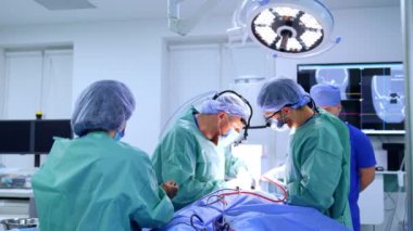 İki cerrah parlak lambanın altında hastanın üzerine eğildi. Kadın hemşire yanında duruyor. Anestezi uzmanı arka planda yürüyor.