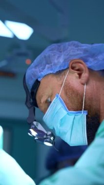 Ameliyat sırasında sakallı beyaz cerrahın konsantre yüzü. Doktor cerrahi loplara bakarak ameliyat yapıyor. Dikey video.