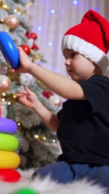 Siyah tişörtlü çocuk oyuncak piramitlerle oynuyor. Noel ağacının yanında Noel Baba şapkası takan bir çocuk. Düşük açı. Dikey video