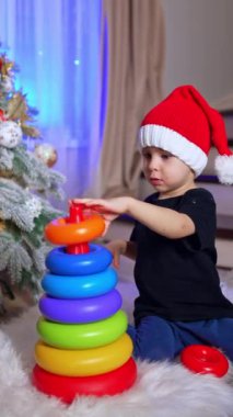 Güzel bebek piramidi birleştiriyor ve sonra onu aşağı itiyor. Noel Baba şapkalı çocuk Noel ağacının yanında oynuyor. Dikey video