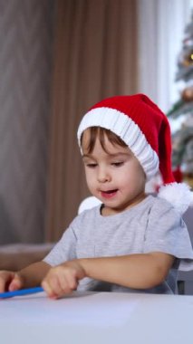 Masada oturan kırmızı şapkalı güzel erkek bebek. Mavi kalemle yeni yürümeye başlayan çocuk çiziyor. Arkaplanda bulanık bir Noel ağacı. Dikey video