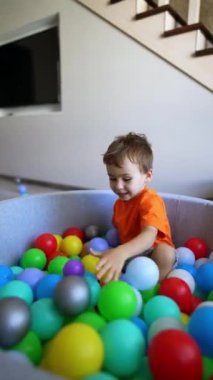 Şirin çocuk kuru havuzda oturuyor. Bebek çanaktan plastik bir top alıyor. Dikey video.