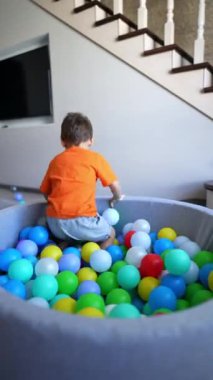 Turuncu tişört giyen neşeli bebek kuru havuzda duruyor. Mutlu çocuk toplarla oynarken sevimli bir şekilde güler. Dikey video.