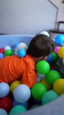 Enerjik erkek bebek kuru havuzda renkli topların içinde yuvarlanıyor. Mutlu çocuk ayağa kalktı ve topları fırlattı. Dikey video.