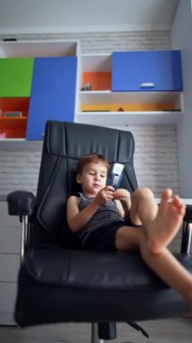 Rahat beyaz erkek bebek büyük siyah deri bir sandalyede oturuyor. Çocuk elinde sürgülü bir telefon tutuyor ve telefonda konuşuyormuş gibi yapıyor. Dikey video.