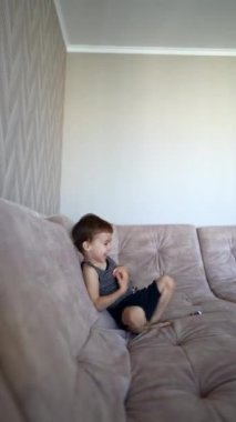 Kafkasyalı erkek bebek büyük kanepede oturuyor. Mutlu çocuk kameraya oyuncak atıyor. Dikey video.