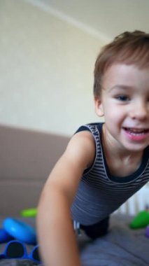Yatakta oturan bir çocuk kameraya yaklaşıyor. Sevimli erkek bebek oyuncağı yatağın kenarına itiyor. Dikey video.