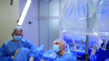 Tezgahta iki plastik poşet ve tuz çözeltisi asılı duruyor. Kapatın. Erkek doktor ve yardımcı hemşire bulanık arka planda çalışıyor..