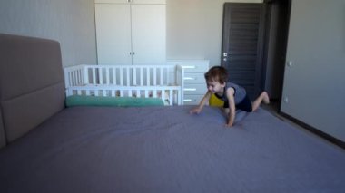 Sevimli gülümseyen küçük çocuk büyük yatağa çıkıyor. Sevimli aktif erkek bebek yatakta zıplamaya başlıyor..