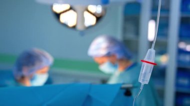 Damardan serum fizyolojik solüsyonlu. Kapatın. Şapka ve maskeli iki doktor bulanık arkaplanda çalışıyor..