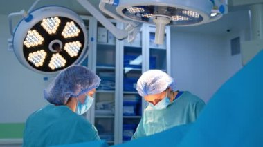 Modern cerrahi odasındaki parlak lambaların altında duran kadın sağlık görevlileri. Cerrah metal bir alet uyguluyor ve hemşire sünger kullanıyor..