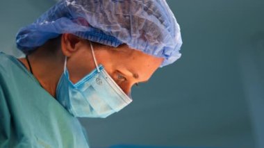 Maskeli ve şapkalı, konsantre olmuş beyaz kadın cerrah aşağıya bakıyor. Doktor metal tıbbi aletler uyguluyor. Kapat..