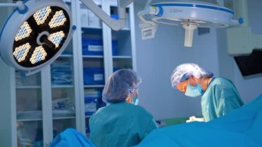 Cerrah ve ameliyat hemşiresi ameliyat masasının üstündeki yuvarlak lambaların altında çalışıyor. Kadın sıhhiyeciler ameliyat yapıyor.