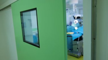 Modern bir ameliyathaneye açılan pencereli bir kapı. Doktorlar ameliyat masasında çalışıyor. Bulanık arkaplan.
