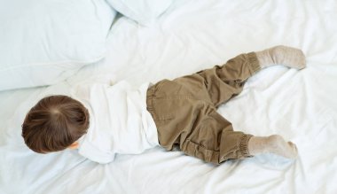 Bebek yumuşak yatakta şen şakrak sürünüyor. Beyaz gömlekli ve kahverengi pantolonlu bir bebek, parlak bir odada yumuşak bir yatak ile rahat bir yatakta emekliyor..