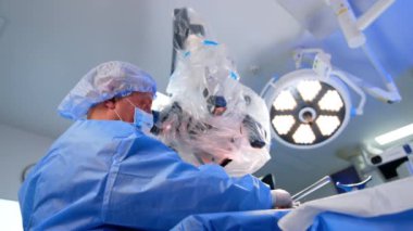 Teknolojik mikroskopta profesyonel erkek cerrah oturuyor. Doktorun ameliyat masasında araç seçerken düşük açılı görüntüsü. Hemşire aletleri beyin cerrahına verir..