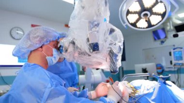 Şapkalı ve maskeli beyaz erkek cerrah mikroskoba bakar ve elektrikli alet kullanır. Beyin cerrahı ameliyatta. Yan görünüm.