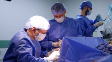 Deneyimli erkek cerrahlar beyin ameliyatı yaparlar. Arkaplandaki sıhhiye, nöroşirürjide kullanılacak mikroskop hazırlıyor..