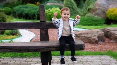 Güzel beyaz bir erkek bebek tahta bankta oturuyor. Sevimli çocuk ellerini sallıyor ve gülümsüyor..