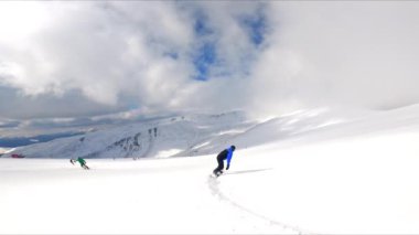 Dağlarda snowboard yapan bir sporcuyu takip ediyorum. Arka planda bulutlu gökyüzü altında büyüleyici manzara.