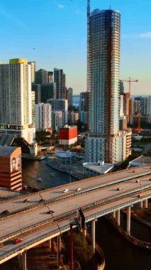 Miami şehir merkezindeki otobanlardan arabalar hızla geçiyor. Gün batımında tepeden güneşli şehir manzarası. Dikey video