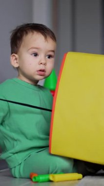 Beyaz çocuk sarı oyuncak kutusunun yanında oturuyor ve siyah kabloyla oynuyor. Sevimli bebek şaşırmış bir şekilde kameraya bakıyor. Dikey video