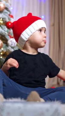 Güzel beyaz çocuk Noel ağacının yanında yerde oturuyor. Kırmızı şapkalı çocuk ayaklarını halıya vuruyor. Düşük açı. Dikey video