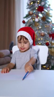 Mavi kalemle kırmızı Noel Baba şapkası çizen odaklanmış bebek. Yeni yürümeye başlayan çocuk Noel tatilinde eğleniyor. Dikey video