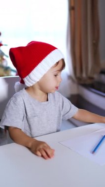 Kırmızı şapkalı sevimli erkek bebek elinde kalemle masadan kalkar. Çocuk masanın arkasına saklanarak yere oturuyor. Dikey video