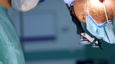 Maskeli, koruyucu gözlüklü ve şapkalı beyaz bir cerrahın sakallı yüzü dikkatle aşağıya bakıyor. Kapatın. Doktor çalışıyor..
