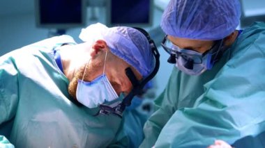 Kızıl saçlı sakallı cerrah hastanın üzerine eğildi. Cerrahi loupes 'taki doktorlar ameliyatta işbirliği yapıyorlar..
