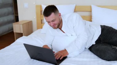 Beyaz gömlek ve siyah kot giyen beyaz sakallı beyaz adam yatağın üzerinde yatıyor. Adam dizüstü bilgisayar kullanıyor..