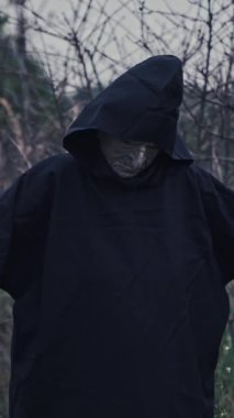 Kapüşonlu, koyu renk kostümlü korkunç adam. Korkunç bir hayaletin portresi. Kolları doğa arka planında. Cadılar Bayramı konsepti Dikey video