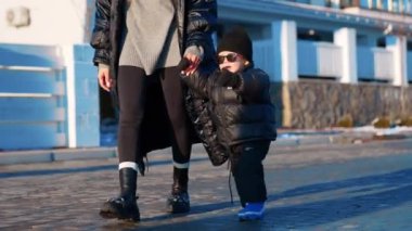 Anne, sevimli çocuğunu elinden tutarak yürütür. Kalın giysiler ve güneş gözlüğü takan yakışıklı bir erkek bebek..