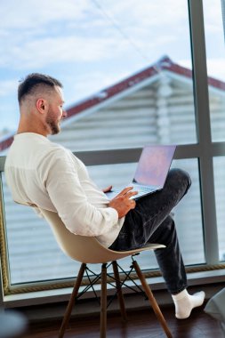 Evdeki pencerenin önünde dizüstü bilgisayarda çalışan adam. Bir adam rahat bir şekilde bir sandalyede oturur ve pencerenin dışındaki manzaranın tadını çıkarırken dizüstü bilgisayarına odaklanır..