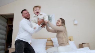 Babası kanepede el sallıyor ve neşeli küçük oğlunu fırlatıyor. Sevgi dolu ebeveynler sevimli oğullarını yanağından öperler. Düşük açı görünümü.