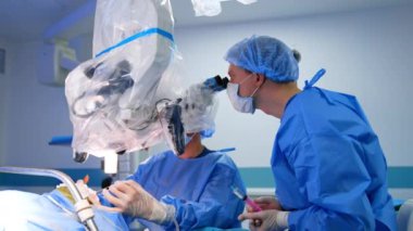 Erkek doktor, mikroskopta sıvı dolu bir şırınga tutuyor. Mikroskobun arkasında oturan ana beyin cerrahı elektrikli aletleri uyguluyor..