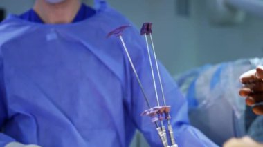 Kahverengi lateks eldivenli cerrahların elleri hastanın vücuduna yerleştirilen uzun metal aletleri düzenler. Mavi cüppeli tanınmayan doktor arka planda duruyor..