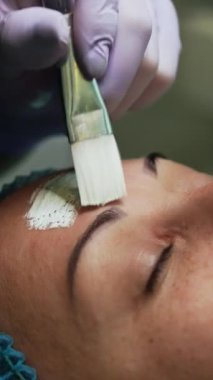 Lastik eldivenli güzellik uzmanı, müşterinin yüzünü maskeyle kaplamak için fırça kullanıyor. Güzellik salonunda gençleştirme prosedürleri. Kapatın. Dikey video