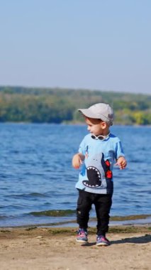 Güneşli nehir sahillerinde eğleniyoruz. Kumsalda oynayan küçük bir çocuk. Dikey video